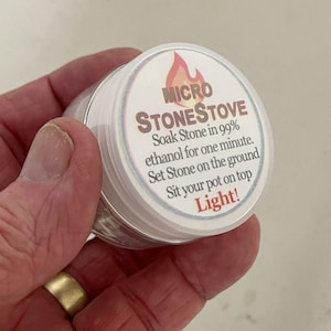 Könnte beinhalten: Ein kleiner, runder Behälter mit weißem Deckel und silbernem Rand. Der Deckel trägt den Text "MICRO STONESTOVE" und Gebrauchsanweisungen. Der Behälter wird in einer Hand gehalten.