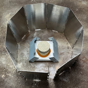 May include: A portable camping stove with a silver wind shield. The stove has a gray base with a circular, layered fuel source. The wind shield is made of multiple hinged panels, creating a protective barrier around the stove.