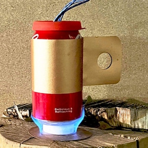Könnte beinhalten: Ein rot-goldenes Objekt in Dosenform mit rotem Deckel und blauer Schnur. Die Dose hat ein goldenes Band und die Worte "Delicious & Refreshing". Es steht auf einer blauen Flammenbasis und ruht auf einer Holzoberfläche.