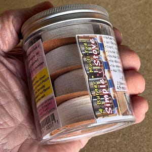 May include: A clear jar with a silver lid containing four white fuel cells with orange bands. A label on the jar reads "4-Burner Multi-fuel Simple Stove". The jar is held in a hand.