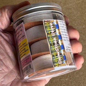 May include: A clear plastic jar containing three round, light-coloured stone discs with orange edges. The label reads SnowStone Stove, 4-Burner, Simmers & Boils, and includes instructions for using bio ethanol fuel. The jar has a silver screw-top lid.