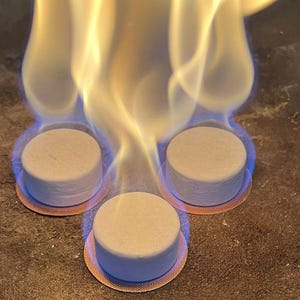 Könnte beinhalten: Drei weiße Brennstofftabletten brennen mit einer blauen und gelben Flamme. Die Tabletten befinden sich auf einem blauen und weißen Untergrund.