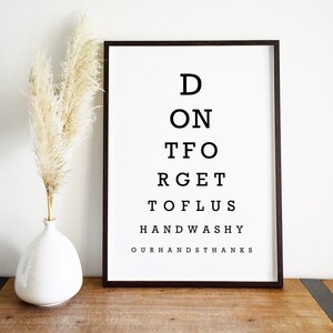 May include: A black framed print with white text that reads "D ON T FO RGET TO FLUS HANDWASHY OURHANDS THANKS". The print is on a wooden table with a white vase with dried pampas grass.