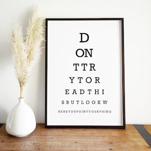 May include: Black and white eye chart print with the text "Don't try to read this but look where you point your thing". The print is framed in a black frame and is sitting on a wooden table.