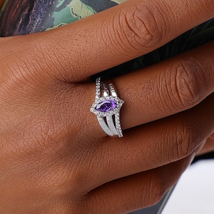 May include: A silver-toned ring featuring a marquise-cut purple gemstone at the center, surrounded by small clear stones. The band is split into two sections, with one side engraved with the year 2021 and the other side featuring clear stones.