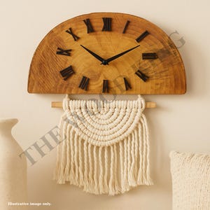 May include: A wooden wall clock in a half-circle shape, featuring black Roman numerals and hands. Beneath the clock hangs a cream macrame wall decoration with a fringe. The clock is mounted on a light-coloured wall.
