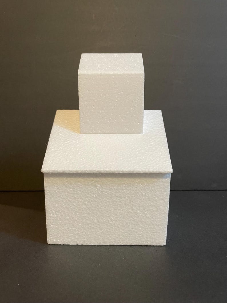 May include: Two white foam cake dummies stacked on top of each other. The bottom dummy is larger than the top dummy.