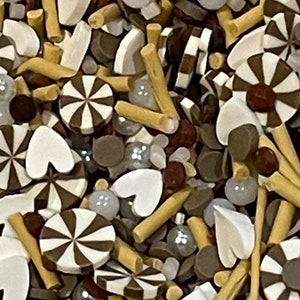 May include: A close-up of a variety of sprinkles in brown, white, and gold. The sprinkles are in the shape of hearts, swirls, and sticks.