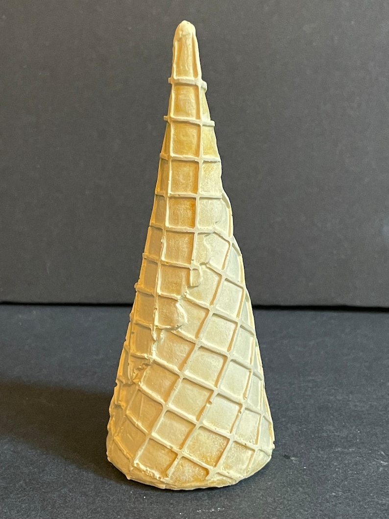 May include: A white waffle cone made of paper, with a grid pattern. The cone is empty and stands upright on a dark surface.