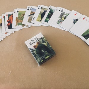 May include: A deck of playing cards with illustrations of extinct animals. The cards are arranged in a semi-circle with the box of cards in the center. The box is white with black text that reads "The 6th Extinction".