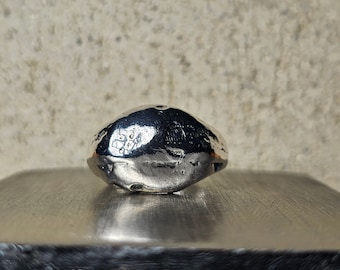 May include: A silver signet ring with a textured, rounded surface. The ring is sitting on a gray surface.