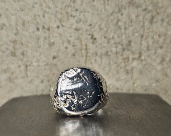 May include: A silver ring with a textured, round face. The ring is sitting on a black surface.
