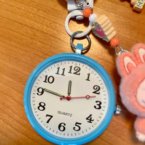 Könnte beinhalten: Eine hellblaue Quarzuhr mit weißem Zifferblatt und schwarzen Zahlen. Die Uhr ist an einem Schlüsselanhänger mit orangefarbenen und weißen Akzenten und einem rosa Hasenanhänger befestigt. Die Uhrzeit ist ungefähr 1:00 Uhr.