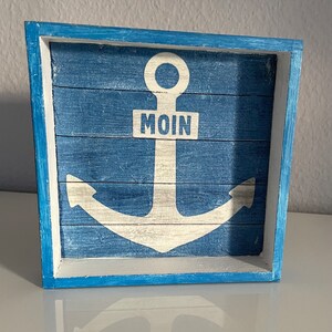 Könnte beinhalten: Ein blaues Holzschild mit einem weißen Anker und dem Wort "MOIN" in der Mitte. Das Schild hat einen verwitterten Look, als wäre es vom Meer gezeichnet worden.