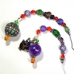 May include: A beaded ornament with a variety of colorful beads, including purple, green, red, and brown. The ornament features a charm of a camel and a large, textured purple bead. The beads are strung together with a thin, white string.