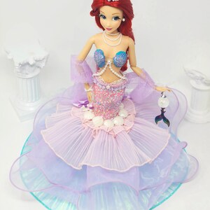 May include: A doll of a mermaid with red hair, wearing a tiara, pearl necklace, and a seashell bra. The doll's skirt is layered with pink, purple, and blue fabrics. A small, silver tail charm hangs from her hand.