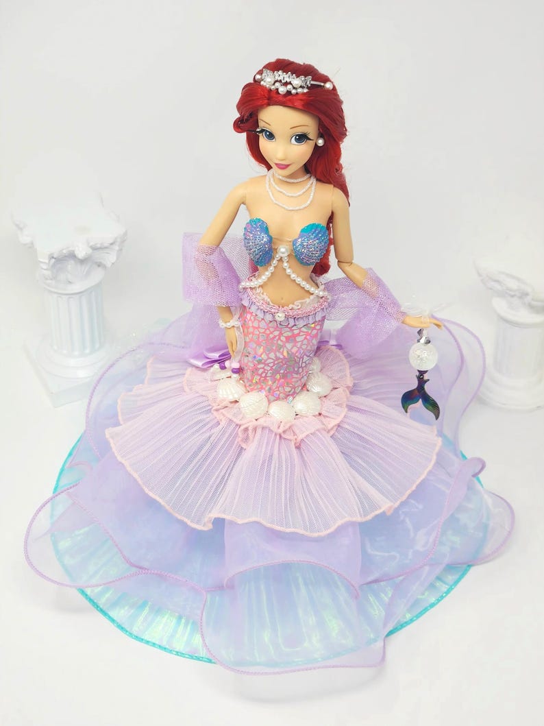 May include: A doll of a mermaid with red hair, wearing a tiara, pearl necklace, and a seashell bra. The doll's skirt is layered with pink, purple, and blue fabrics. A small, silver tail charm hangs from her hand.