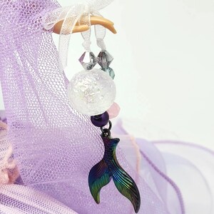 May include: A whimsical charm featuring a mermaid tail pendant with iridescent colors of green, blue, and purple. The charm is adorned with a clear, spherical bead, pink and purple beads, and clear faceted beads, suspended from a white ribbon.