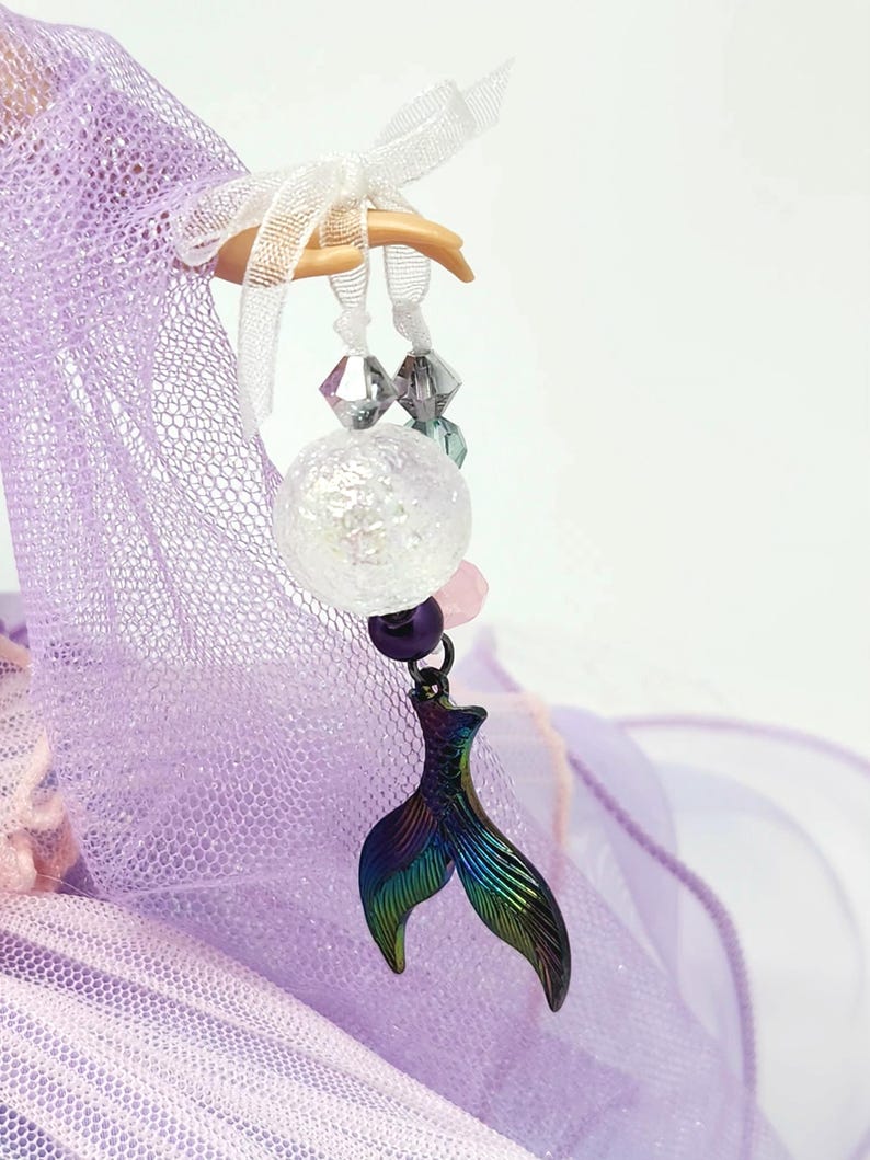 May include: A whimsical charm featuring a mermaid tail pendant with iridescent colors of green, blue, and purple. The charm is adorned with a clear, spherical bead, pink and purple beads, and clear faceted beads, suspended from a white ribbon.
