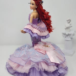 Op de afbeelding: Een pop met lang rood haar en een gedetailleerde paarse, roze en blauwe jurk. De pop heeft een parelketting en een versierd hoofddeksel. De jurk heeft lagen stof en een grote strik met een versierd middelpunt.
