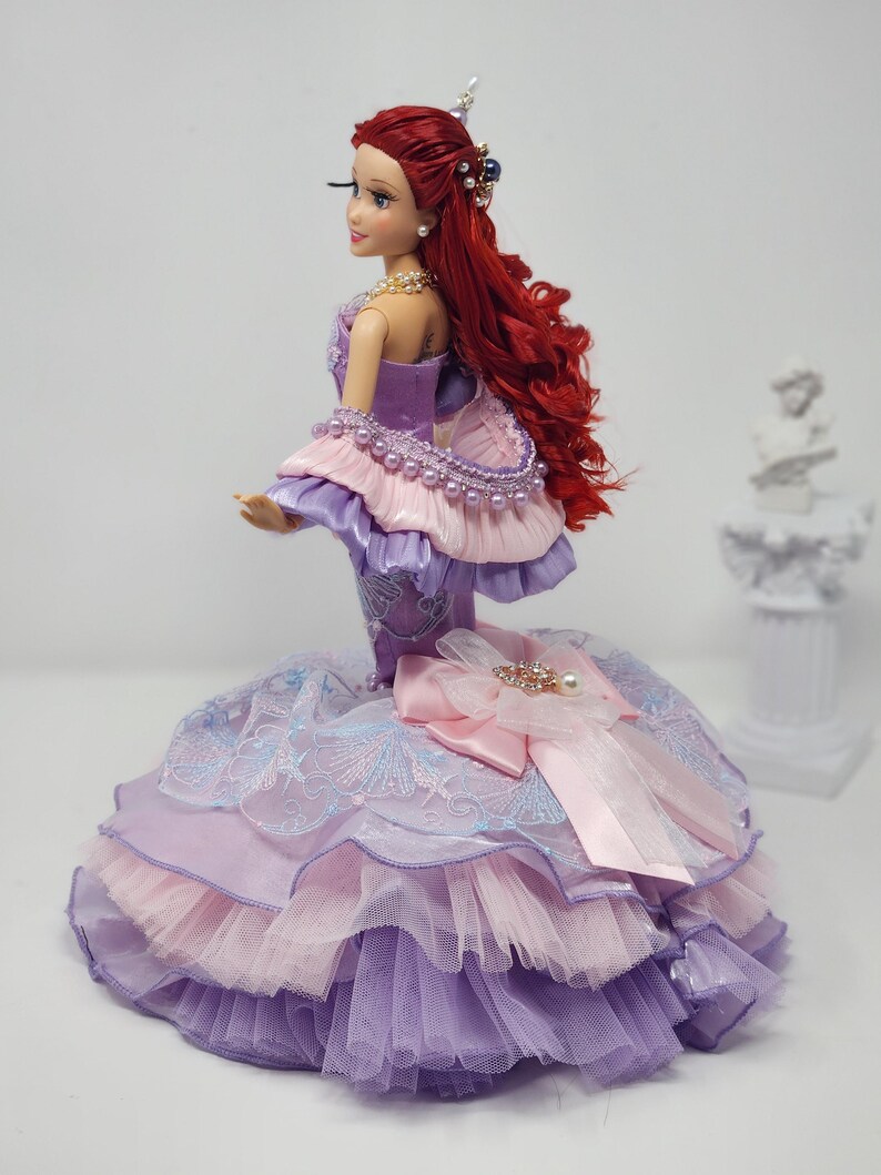 Op de afbeelding: Een pop met lang rood haar en een gedetailleerde paarse, roze en blauwe jurk. De pop heeft een parelketting en een versierd hoofddeksel. De jurk heeft lagen stof en een grote strik met een versierd middelpunt.