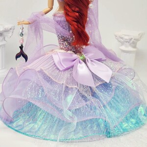 May include: A doll dressed as a mermaid, featuring a red wig and a lavender and blue gown. The gown has a large bow and is adorned with shimmering fabric. A tail-shaped charm hangs from the doll's hand, adding to the mermaid theme.