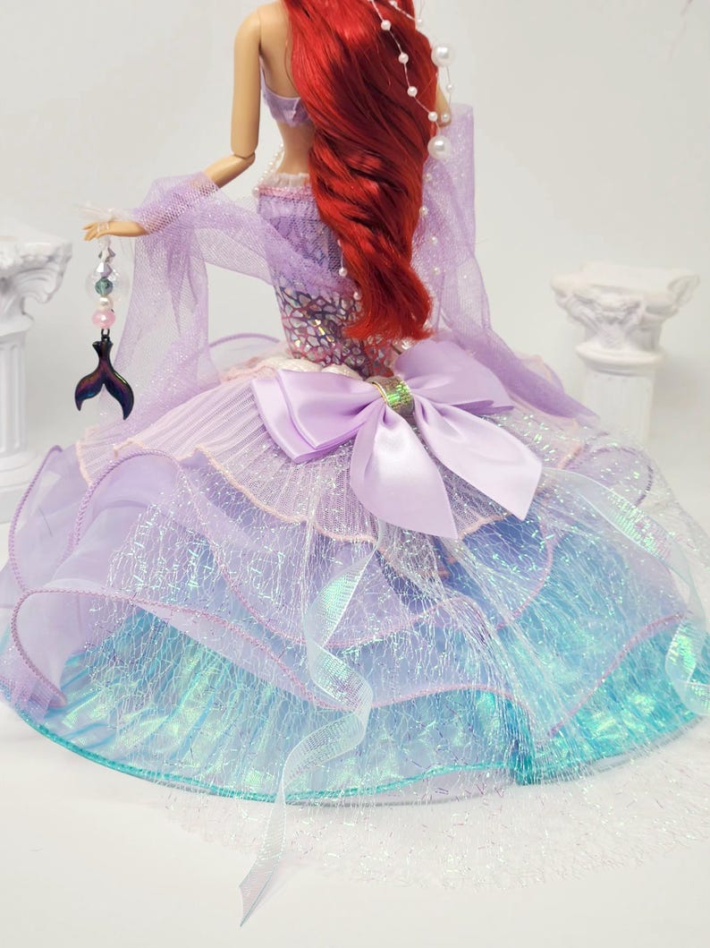 May include: A doll dressed as a mermaid, featuring a red wig and a lavender and blue gown. The gown has a large bow and is adorned with shimmering fabric. A tail-shaped charm hangs from the doll's hand, adding to the mermaid theme.