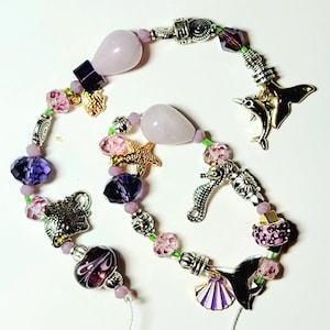 May include: A handmade beaded necklace featuring a variety of charms and beads. The necklace includes light purple teardrop-shaped stones, faceted purple beads, and silver-toned charms such as a seahorse, dolphin, and starfish. Pink and purple beads are strung on a green cord.