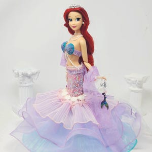 May include: A doll of a mermaid with long red hair, wearing a seashell bra and a layered purple and blue skirt. The doll has a silver tiara and a pearl necklace. The mermaid is holding a small, clear purse with a mermaid tail charm.