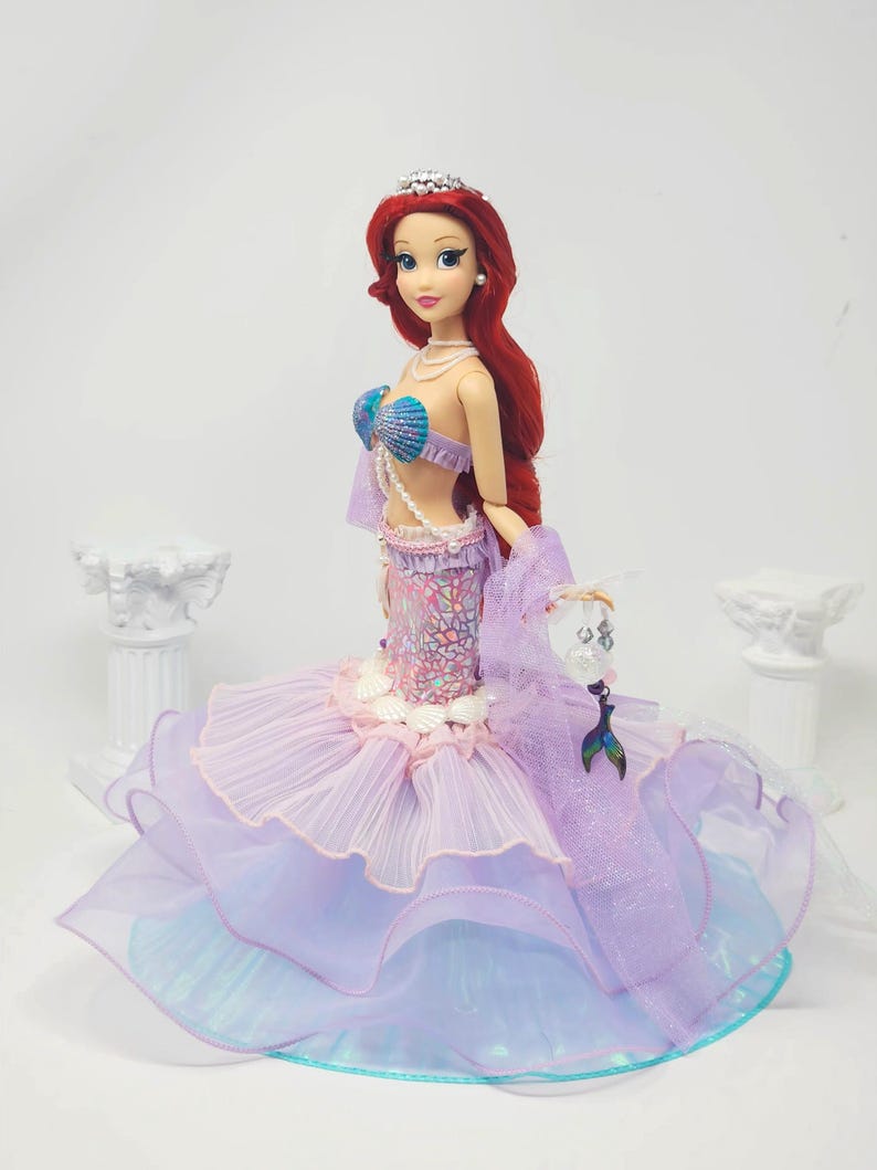 May include: A doll of a mermaid with long red hair, wearing a seashell bra and a layered purple and blue skirt. The doll has a silver tiara and a pearl necklace. The mermaid is holding a small, clear purse with a mermaid tail charm.