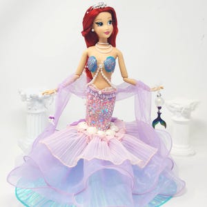 May include: A doll dressed as a mermaid, featuring a red-haired doll wearing a tiara, shell-shaped top, and a layered, ruffled skirt in shades of purple and blue. The doll holds a decorative tail-shaped charm.
