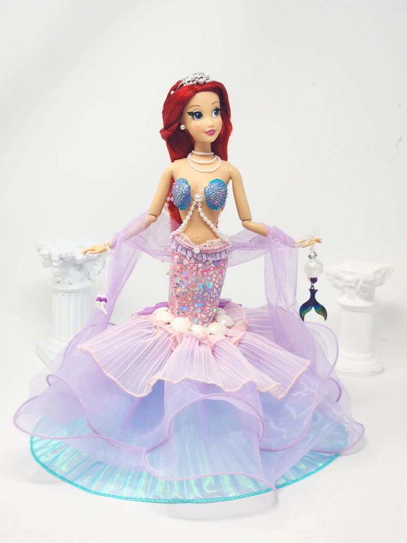 May include: A doll dressed as a mermaid, featuring a red-haired doll wearing a tiara, shell-shaped top, and a layered, ruffled skirt in shades of purple and blue. The doll holds a decorative tail-shaped charm.