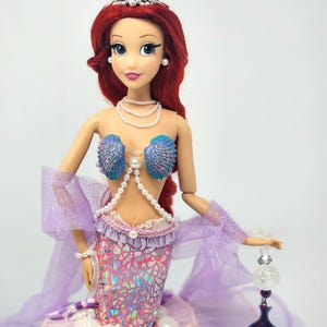 May include: A doll dressed as a mermaid, featuring red hair, a pearl necklace, and a seashell top. The doll's tail is a shimmering pink and purple, with a layered tulle skirt. A silver tiara and a beaded accessory complete the look.