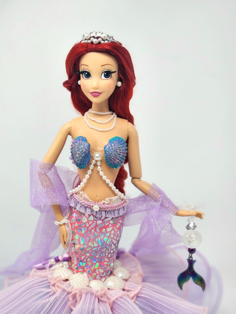 May include: A doll dressed as a mermaid, featuring red hair, a pearl necklace, and a seashell top. The doll's tail is a shimmering pink and purple, with a layered tulle skirt. A silver tiara and a beaded accessory complete the look.