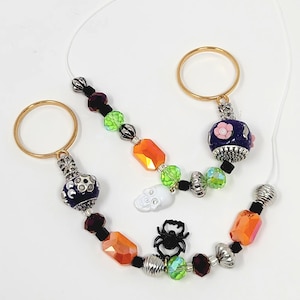May include: Three Halloween-themed beaded charms with gold-coloured rings. The charms feature orange, green, black, and purple beads, with skull and spider accents. The charms are strung on a thin, white cord.