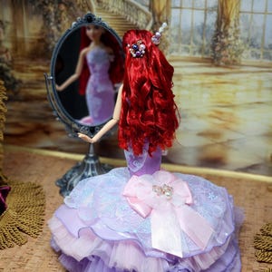 Op de afbeelding: Een pop met lang, levendig rood haar en een lavendel- en roze jurk. De pop staat voor een spiegel en weerspiegelt haar beeld. De jurk heeft een grote roze strik en een gelaagde tule rok. Het haar van de pop is versierd met decoratieve haarspelden.