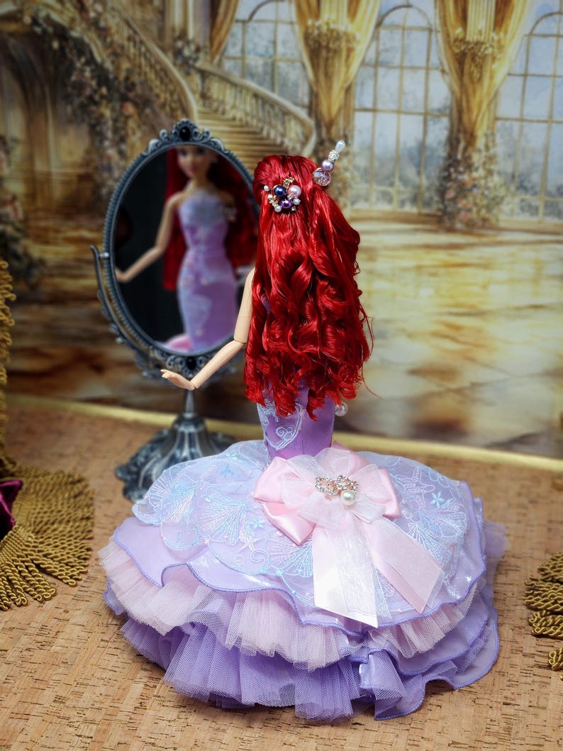 Op de afbeelding: Een pop met lang, levendig rood haar en een lavendel- en roze jurk. De pop staat voor een spiegel en weerspiegelt haar beeld. De jurk heeft een grote roze strik en een gelaagde tule rok. Het haar van de pop is versierd met decoratieve haarspelden.