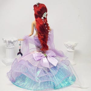 May include: A doll with long red hair, wearing a purple and blue gown. The gown features a large bow and layered skirt. The doll has a tiara and pearl accents in her hair. A decorative tail charm hangs from her hand.