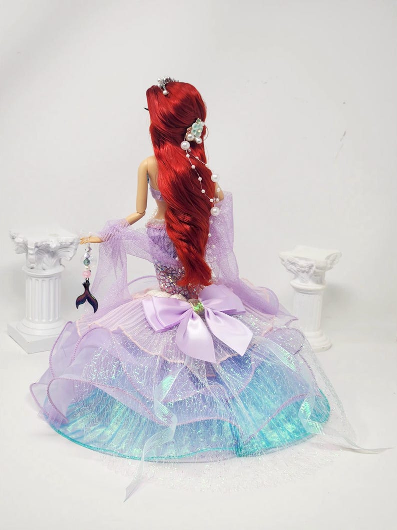 May include: A doll with long red hair, wearing a purple and blue gown. The gown features a large bow and layered skirt. The doll has a tiara and pearl accents in her hair. A decorative tail charm hangs from her hand.
