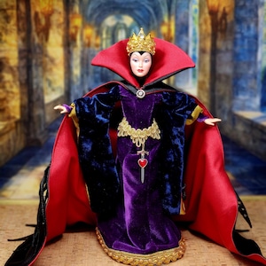 May include: A doll dressed as a queen, wearing a gold crown, red cape, and purple velvet gown with gold trim. The doll has a stern expression and is posed with arms outstretched. A silver pendant with a red heart hangs from the waist.
