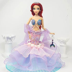 May include: A doll dressed as a mermaid, featuring a red wig, a silver tiara, and a pearl necklace. The doll's bodice is adorned with blue seashell-shaped cups and a pearl strand. The mermaid's tail is a layered skirt in shades of pink, purple, and blue.