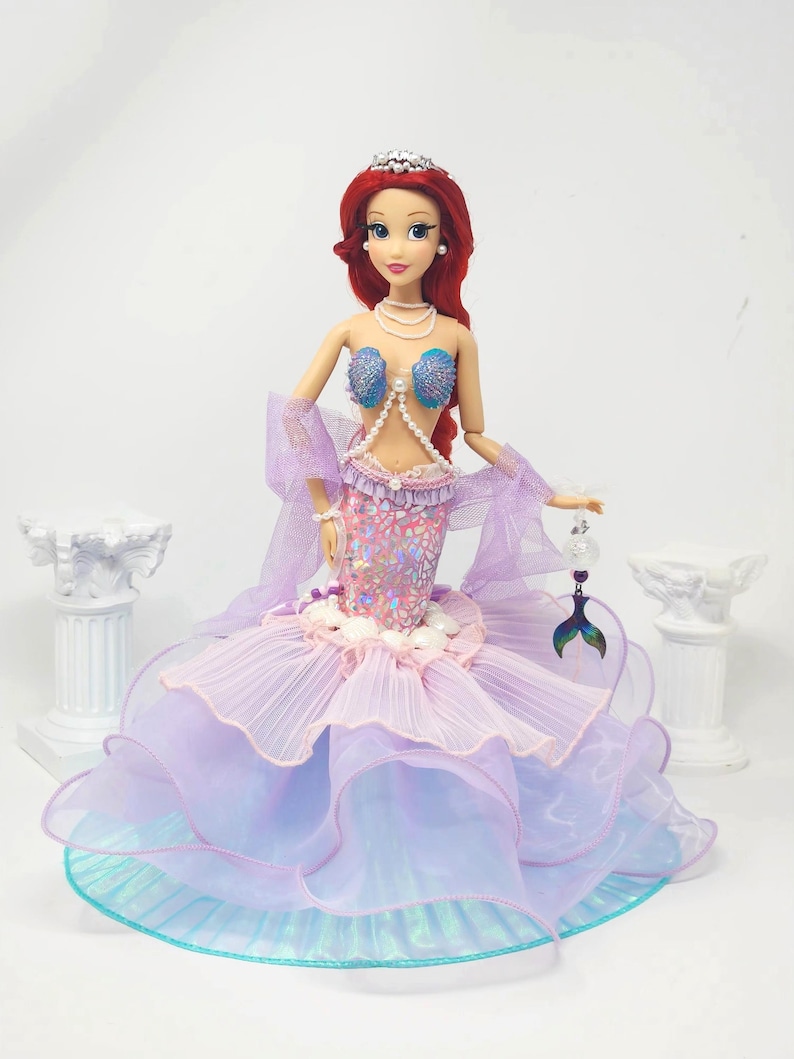 May include: A doll dressed as a mermaid, featuring a red wig, a silver tiara, and a pearl necklace. The doll's bodice is adorned with blue seashell-shaped cups and a pearl strand. The mermaid's tail is a layered skirt in shades of pink, purple, and blue.