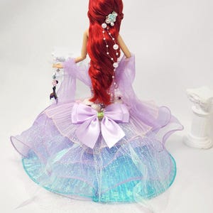 May include: A doll with long, flowing red hair, wearing a lavender and blue dress with a large bow. The doll has a tiara and pearl hair accessories. The dress has a layered, shimmering skirt.