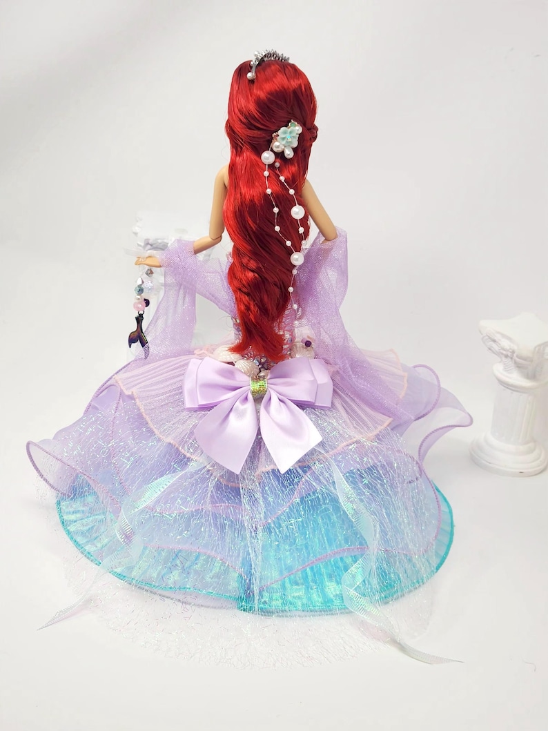 May include: A doll with long, flowing red hair, wearing a lavender and blue dress with a large bow. The doll has a tiara and pearl hair accessories. The dress has a layered, shimmering skirt.