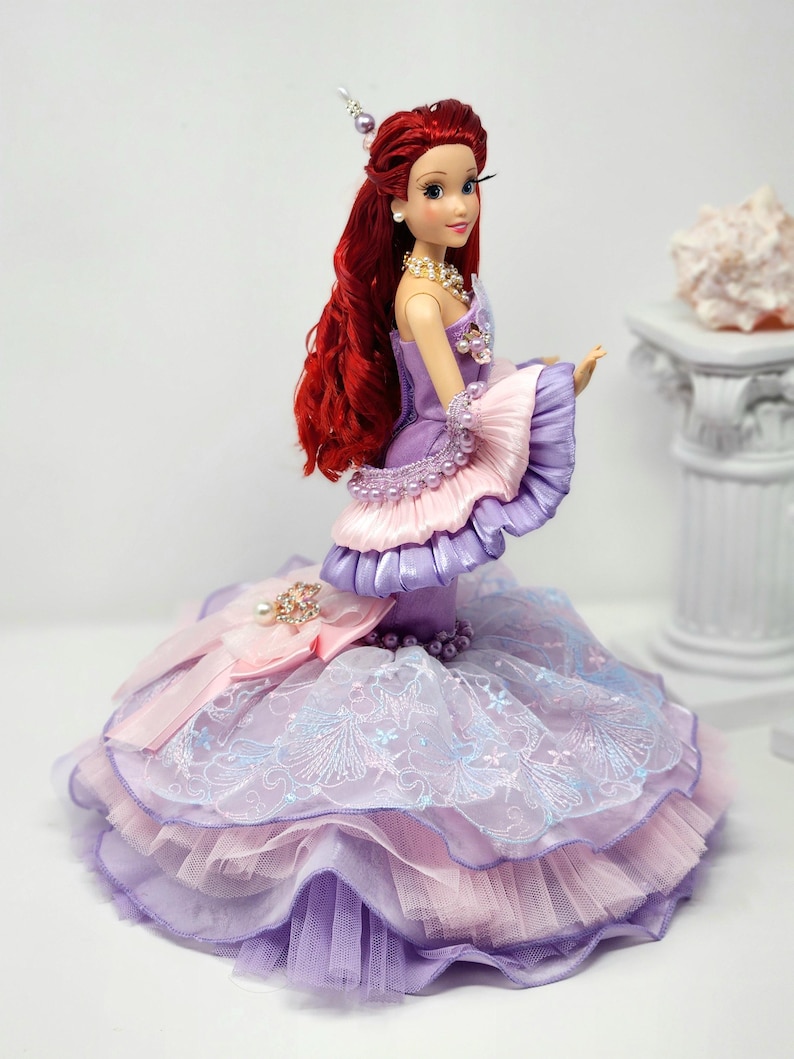 Op de afbeelding: Een pop met lang rood haar, gekleed in een gelaagde jurk in tinten lavendel, roze en paars. De jurk heeft ingewikkelde kant- en lintdetails, met parelaccenten op het lijfje en de mouwen. De pop heeft een parelketting en oorbellen.