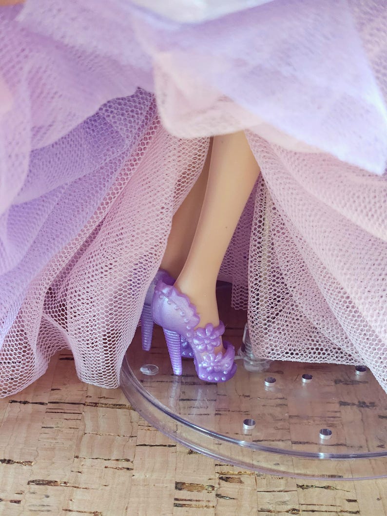 Op de afbeelding: Close-up van de benen van een pop met lavendelkleurige hoge hakken met bloemaccenten. De hakken staan op een doorzichtige standaard en de pop draagt een gelaagde tule jurk in tinten van lavendel en roze.