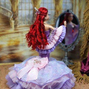 Op de afbeelding: Een pop met lang rood haar en een paarse jurk met een grote roze strik. De pop kijkt in een kleine spiegel. De jurk heeft lagen paarse en roze stof. De pop draagt een parelketting en een sierlijk haaraccessoire.