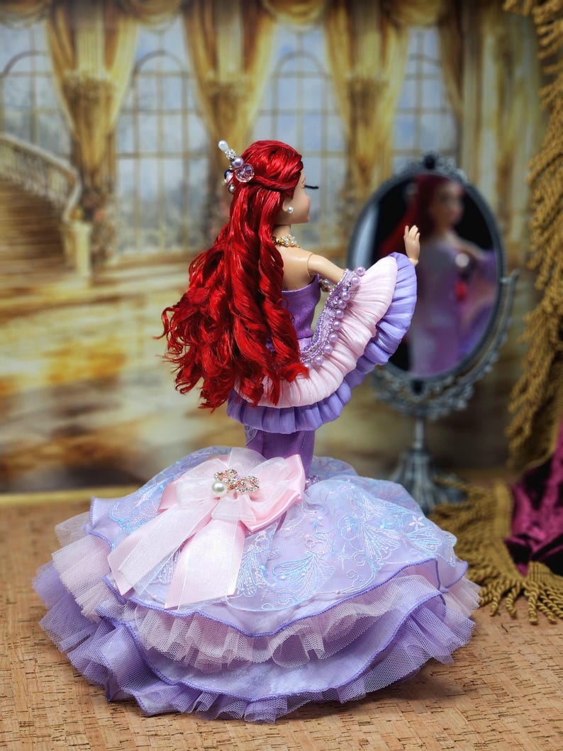 Op de afbeelding: Een pop met lang rood haar en een paarse jurk met een grote roze strik. De pop kijkt in een kleine spiegel. De jurk heeft lagen paarse en roze stof. De pop draagt een parelketting en een sierlijk haaraccessoire.