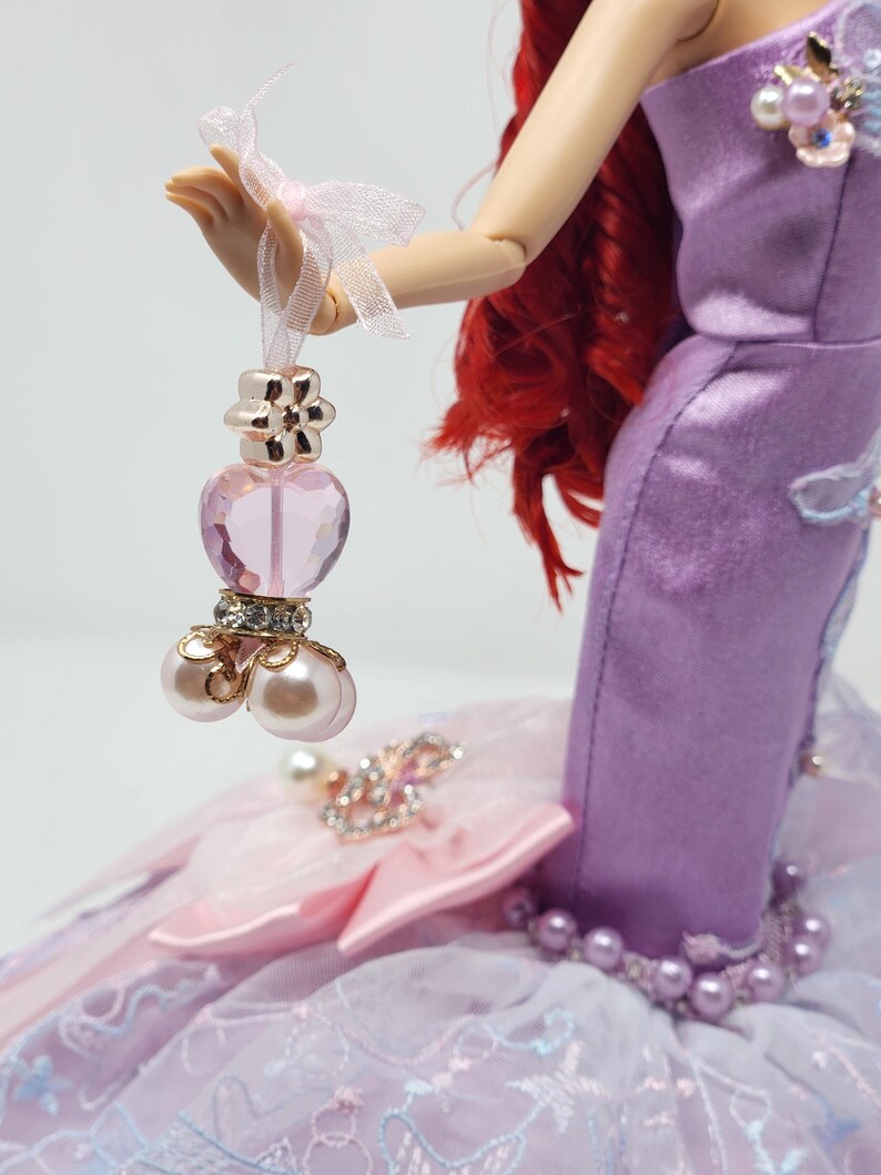 Op de afbeelding: Een pop in een lavendelkleurige jurk versierd met parels en strass. De pop heeft rood haar. Een kleine, hartvormige parfumfles met pareldetails wordt vastgehouden door de hand van de pop.