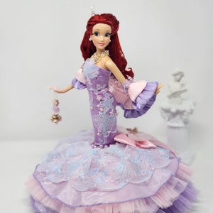Op de afbeelding: Een prinsessenpop met rood haar, gekleed in een lavendelkleurige jurk met gedetailleerde borduursels en een gelaagde rok. De jurk heeft roze en blauwe accenten en de pop is versierd met sieraden.